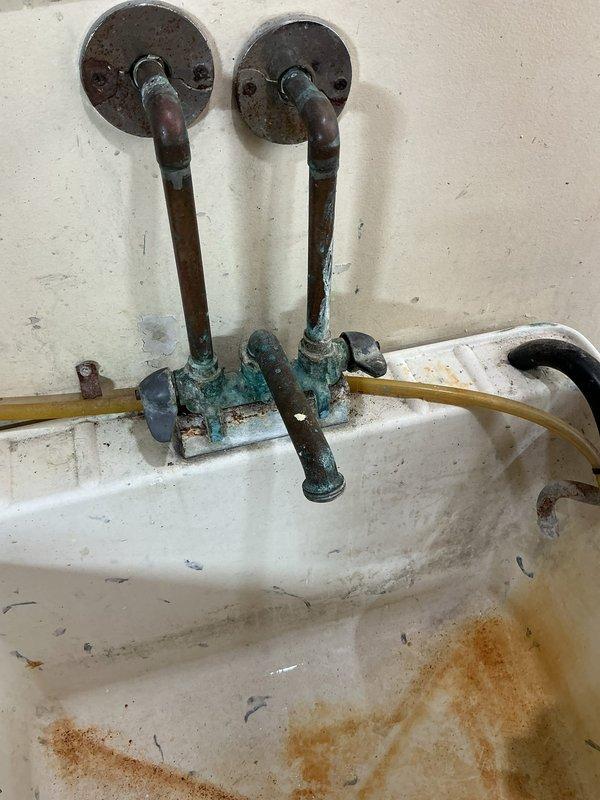 Responded to basement utility sink with dripping faucet complaint. Upon inspection, found severely deteriorated wall-mounted laundry tub faucet assembly with extensive copper oxidation, verdigris buildup on pipes and fittings, and corroded valve components. The vintage plumbing fixtures showed advanced age with compromised seals causing the reported drip. Assessed water supply lines and shutoff valves in ceiling area above fixture. Due to the extensive corrosion throughout the faucet assembly and the obsolete nature of the fixtures, recommended complete replacement of the wall-mounted faucet unit to resolve the leak and prevent future failures.
