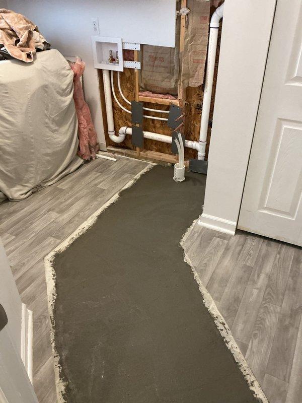 Returned to site to complete cement work following plumbing repair. Applied self-leveling concrete compound to floor area where previous plumbing access was required, creating smooth transition between existing vinyl plank flooring. PVC plumbing lines remain exposed in wall cavity with framing visible, awaiting final wall closure and finishing work.
