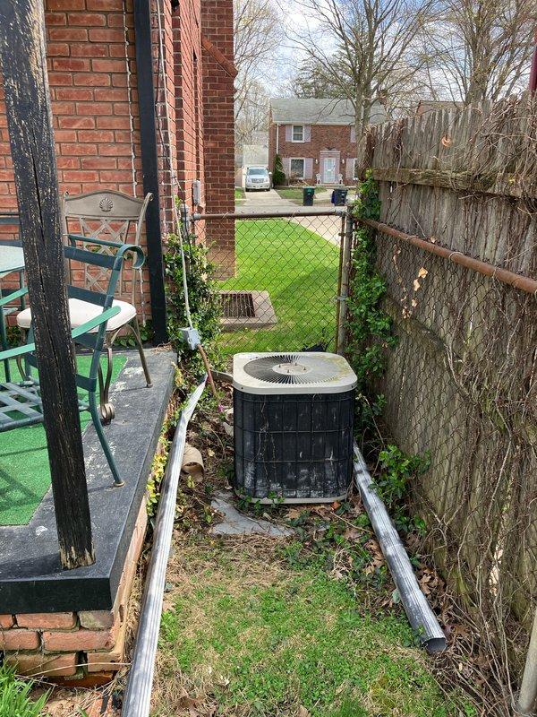 Conducted on-site evaluation for complete HVAC system replacement including outdoor air conditioning unit and basement furnace. Assessed existing cylindrical condenser unit located in narrow side yard between building and chain-link fence, documented equipment condition and installation specifications. Inspected electrical service routing along fence line, evaluated accessibility for replacement equipment, and photographed current system configuration to prepare comprehensive replacement estimate for both cooling and heating systems.