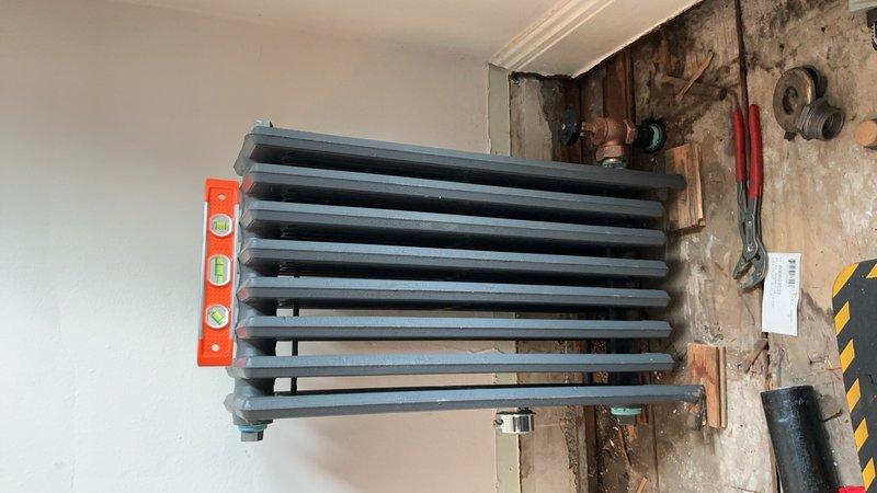 Ordered replacement hydronic radiator unit under PO 1302869-001 with 2-3 week lead time for delivery. Documented existing installation location showing exposed wall framing, copper plumbing connections with shutoff valve, and deteriorated wall conditions requiring radiator replacement. Customer will be contacted upon arrival of new unit for installation scheduling.