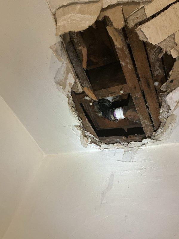 Responded to service call for non-functional hot water tank in basement and bathtub leak in four-family residential building constructed in 1915. Upon inspection, discovered significant water damage requiring access through ceiling to assess plumbing infrastructure and drainage systems. Created access opening in ceiling to expose aged wooden joists and compromised plumbing components. Identified deteriorated piping and water intrusion affecting multiple levels of the structure. Inspected vertical PVC drainage lines running along basement utility area, noting purple primer markings indicating previous installation work. Found evidence of ongoing water damage at wall base with exposed substrate and accumulated debris. Evaluated hot water heater located in basement adjacent to furnace, assessing operational status and connections. Performed comprehensive inspection of both plumbing issues as requested, documenting conditions through photography for repair planning. Damage extent required 