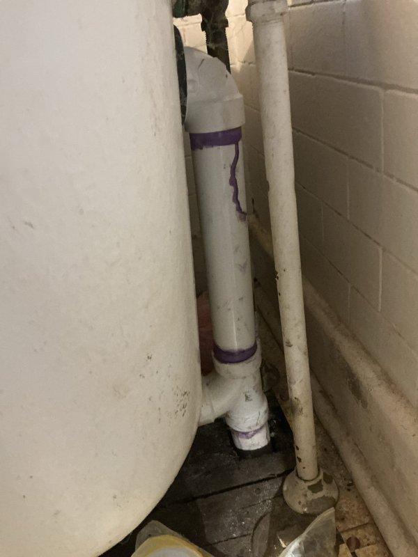 Responded to service call for non-functional hot water tank in basement and bathtub leak in four-family residential building constructed in 1915. Upon inspection, discovered significant water damage requiring access through ceiling to assess plumbing infrastructure and drainage systems. Created access opening in ceiling to expose aged wooden joists and compromised plumbing components. Identified deteriorated piping and water intrusion affecting multiple levels of the structure. Inspected vertical PVC drainage lines running along basement utility area, noting purple primer markings indicating previous installation work. Found evidence of ongoing water damage at wall base with exposed substrate and accumulated debris. Evaluated hot water heater located in basement adjacent to furnace, assessing operational status and connections. Performed comprehensive inspection of both plumbing issues as requested, documenting conditions through photography for repair planning. Damage extent required 