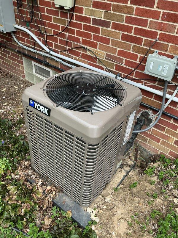 Completed comprehensive Gold AC inspection on 3-year-old York condenser unit with air handler located in basement. Performed multi-point system evaluation including refrigerant line inspection, electrical connection assessment, and condenser coil examination. Unit found operating within normal parameters with proper airflow through louvers and fan assembly functioning correctly. Verified electrical disconnect box and conduit connections secure, inspected copper refrigerant lines and insulation integrity along exterior wall mounting. Documented equipment condition and surrounding area, noting vegetation growth near concrete pad that should be monitored to maintain adequate clearance for optimal airflow and service access.
