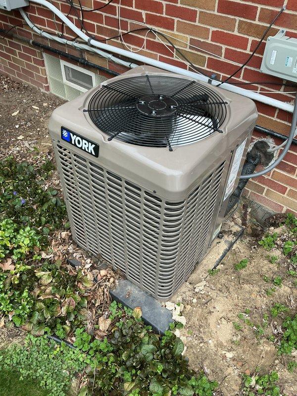 Completed comprehensive Gold AC inspection on 3-year-old York condenser unit with air handler located in basement. Performed multi-point system evaluation including refrigerant line inspection, electrical connection assessment, and condenser coil examination. Unit found operating within normal parameters with proper airflow through louvers and fan assembly functioning correctly. Verified electrical disconnect box and conduit connections secure, inspected copper refrigerant lines and insulation integrity along exterior wall mounting. Documented equipment condition and surrounding area, noting vegetation growth near concrete pad that should be monitored to maintain adequate clearance for optimal airflow and service access.