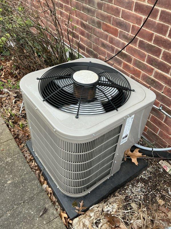 Completed comprehensive Gold AC Inspection and tune-up on 7-year-old residential air conditioning system located on north side of property. Unit is a beige cylindrical condenser with top-discharge fan configuration, mounted on rubber isolation pad against brick exterior wall. Performed full system evaluation including visual inspection of condenser cabinet, coil fins, fan assembly, and electrical connections. Verified proper refrigerant line routing and insulation integrity. Checked condenser pad levelness and stability. Noted presence of seasonal debris including fallen leaves and dried vegetation around unit base and on concrete pad. Inspected manufacturer data plate for model and serial number documentation. Evaluated overall unit condition consistent with 7-8 years of operation. System operational and functioning within normal parameters upon completion of maintenance service.