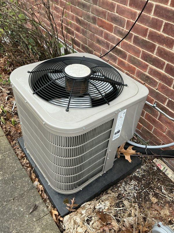 Completed comprehensive Gold AC Inspection and tune-up on 7-year-old residential air conditioning system located on north side of property. Unit is a beige cylindrical condenser with top-discharge fan configuration, mounted on rubber isolation pad against brick exterior wall. Performed full system evaluation including visual inspection of condenser cabinet, coil fins, fan assembly, and electrical connections. Verified proper refrigerant line routing and insulation integrity. Checked condenser pad levelness and stability. Noted presence of seasonal debris including fallen leaves and dried vegetation around unit base and on concrete pad. Inspected manufacturer data plate for model and serial number documentation. Evaluated overall unit condition consistent with 7-8 years of operation. System operational and functioning within normal parameters upon completion of maintenance service.