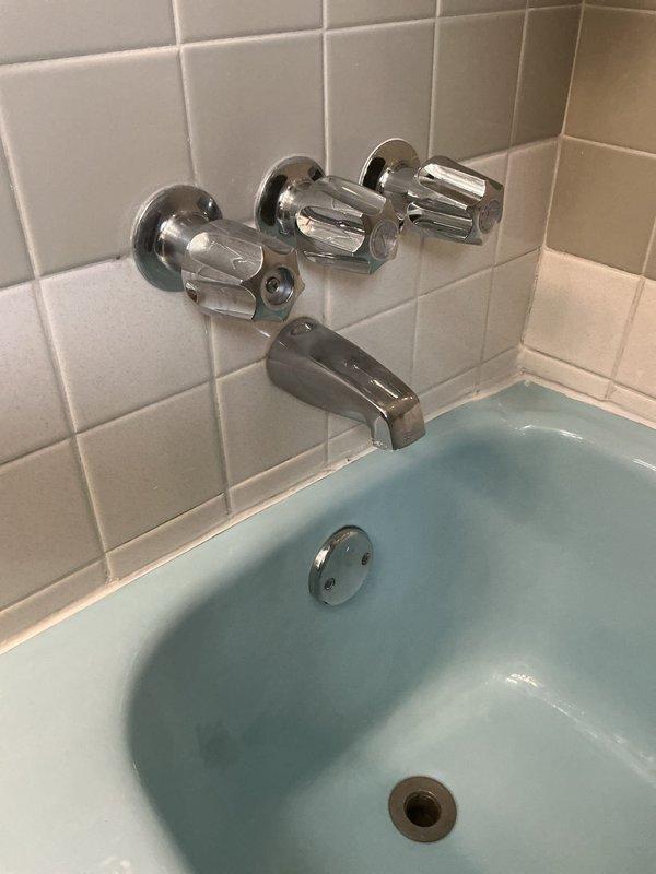 Responded to service call for shower valve conversion from three-valve to single-valve system. Performed diagnostic assessment of existing chrome-finished bathtub faucet assembly with three handles (two cross-handle valves and central diverter) mounted on white tile wall above vintage light blue bathtub. Accessed plumbing infrastructure through wall opening to inspect existing supply lines, including hot and cold water pipes with brass fittings and valves. Evaluated current configuration and measured installation parameters. Provided detailed quote for converting three-valve system to modern single-valve shower control unit, including necessary modifications to existing plumbing connections and wall preparation requirements.