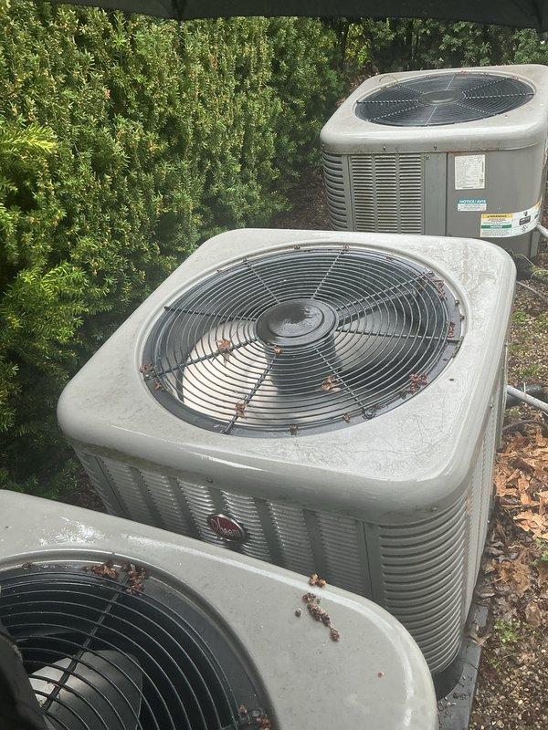 Responded to no cooling complaint on residential property with multiple HVAC systems. Upon arrival, located and inspected three outdoor condenser units positioned on gravel pad with adequate clearance. Units showed signs of normal wear with debris accumulation on top surfaces including leaves and dirt. Verified all condensers were beige-colored cabinet style units with wire fan guards and louvered side panels for proper ventilation. Noted condensate drainage lines present and vegetation growth in close proximity to equipment. Documented equipment condition through photography showing manufacturer labels and warning stickers visible on unit panels. All units appeared to be standard residential split-system configurations with top-discharge fan assemblies mounted on concrete pads with gravel surround for drainage.