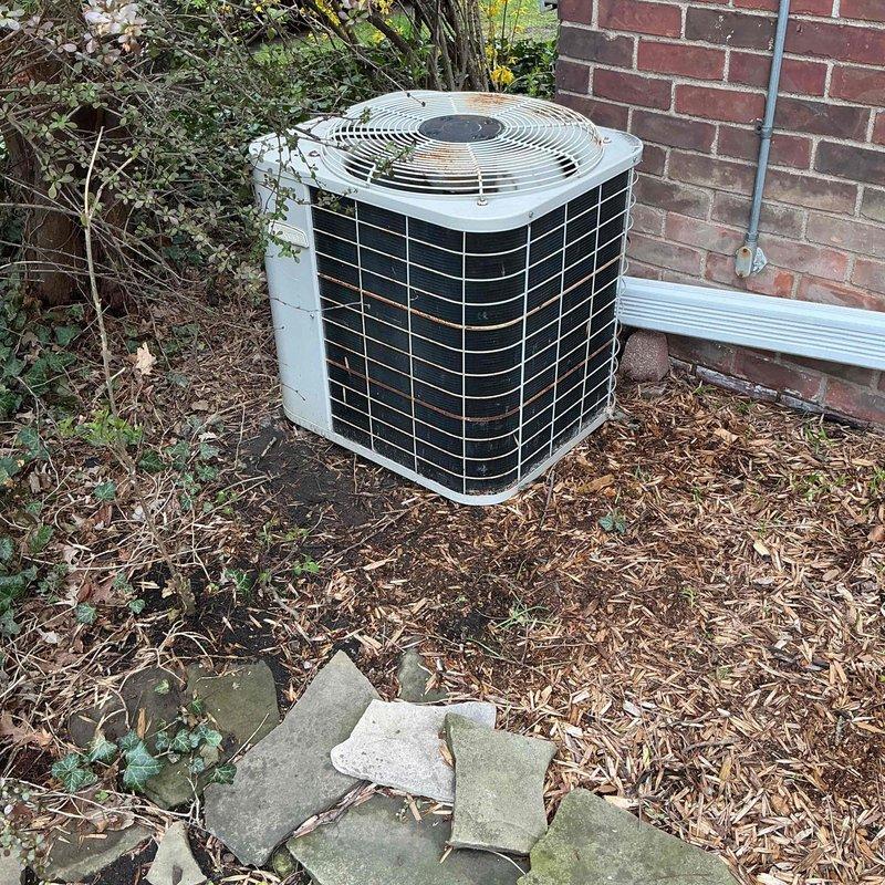 Completed comprehensive Gold AC inspection on very old residential air conditioning system with air handler located in basement. Outdoor condenser unit showed significant age-related wear including rust staining, discoloration, and weathering on exterior casing and mounting hardware. Noted debris accumulation around unit including leaves, twigs, and organic matter in surrounding mulch bed, with overgrown vegetation encroaching near equipment requiring clearance for proper airflow and system efficiency.