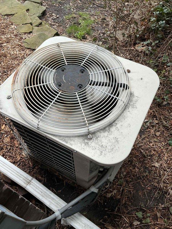 Completed comprehensive Gold AC inspection on very old residential air conditioning system with air handler located in basement. Outdoor condenser unit showed significant age-related wear including rust staining, discoloration, and weathering on exterior casing and mounting hardware. Noted debris accumulation around unit including leaves, twigs, and organic matter in surrounding mulch bed, with overgrown vegetation encroaching near equipment requiring clearance for proper airflow and system efficiency.