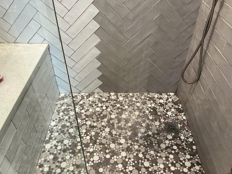 Responded to water leak originating from shower area and penetrating into adjacent bedroom. Upon inspection, identified active water intrusion affecting ceiling or wall cavity between tiled shower enclosure and daughter's bedroom. Documented conditions of modern herringbone-patterned tile shower with pebble floor mosaic and visible water damage evidence in carpeted bedroom area including moisture staining at wall-floor junction. Assessed leak source and provided recommendations for repair of compromised waterproofing membrane or plumbing fixture connection to prevent further water migration into living space.