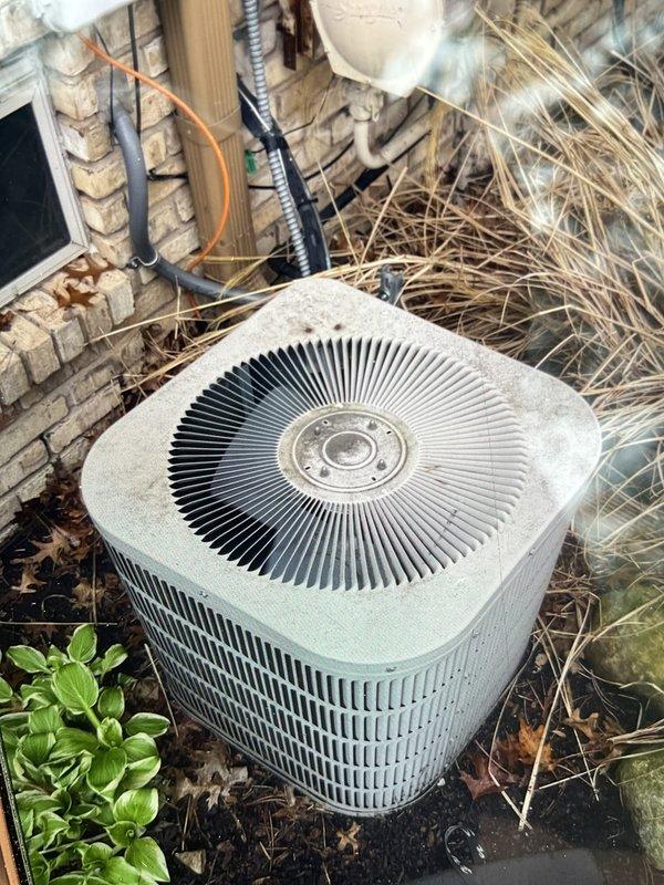 Completed comprehensive Gold AC inspection on residential split system with air handler located in basement and outdoor condenser unit. Inspected triangular air filter showing moderate dust accumulation with pleated media intact and functional. Evaluated outdoor condenser unit positioned near building foundation, noting significant debris accumulation including dried vegetation, straw, and organic matter surrounding unit requiring cleanup for optimal airflow and system efficiency. Documented equipment condition through photographic inspection, verified operational status of approximately 20-year-old system, and assessed accessibility of components for future maintenance requirements.