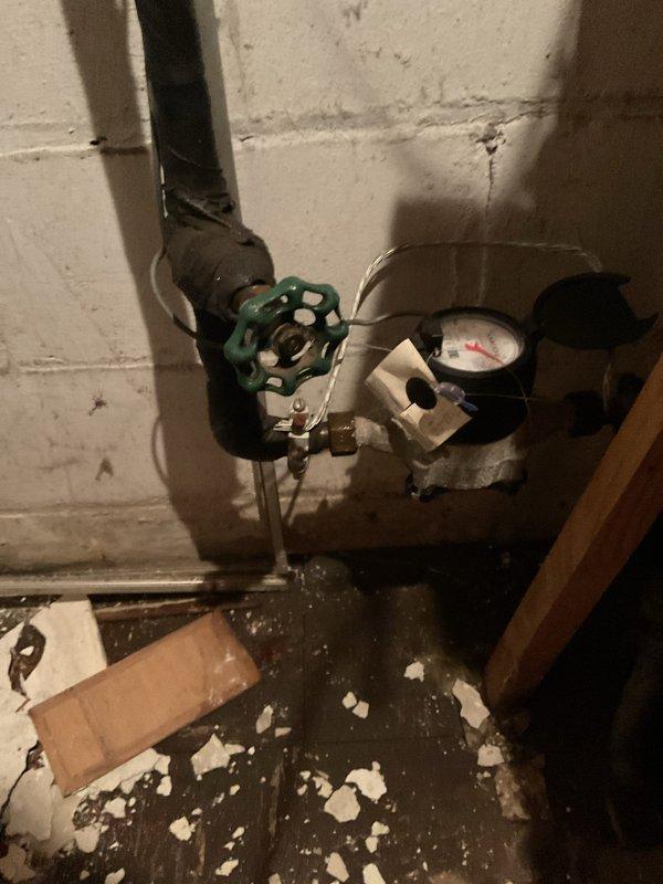 Responded to leaking main waterline in basement of century-old residential property. Upon inspection, identified active leak at vertical pipe penetration point where main water supply line emerges from basement floor. The compromised section exhibited deteriorated insulation wrapping and visible water seepage around pipe-to-floor junction. Shut off water supply at main gate valve near meter assembly to stop active leak. Assessed copper piping system and water meter installation mounted on concrete block foundation wall. Documented existing conditions including aged plumbing infrastructure, corroded pipe coverings, and water damage to surrounding concrete floor area. Recommended complete replacement of affected main line section due to age-related deterioration and potential for future failure in adjacent piping.