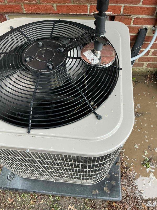 Completed comprehensive Gold AC inspection on 9-year-old residential HVAC system with furnace located in basement. Performed thorough evaluation of outdoor condensing unit positioned on concrete pad against exterior brick wall, confirming operational status of fan motor assembly, capacitor, and electrical connections. Inspected condenser coil protection mesh, verified proper airflow through intake panels, and assessed electrical conduit routing to junction box mounted on building exterior. Documented unit condition with photographic evidence showing functional components, weathered concrete pad with minor debris accumulation, and PVC drainage line installation. Confirmed single-unit residential system is operating as designed with homeowner present during inspection.