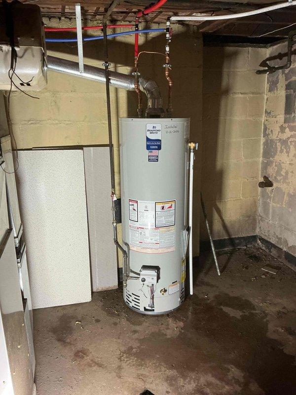 Completed installation of a 40-gallon residential gas-fired water heater in basement utility area. Unit was positioned on concrete floor against cinder block wall with all plumbing connections made using copper piping at inlet and outlet points, brass fittings and valves installed at top connections, and gas line reconnected per code requirements. All manufacturer labels and safety information remain visible and intact on exterior of unit.