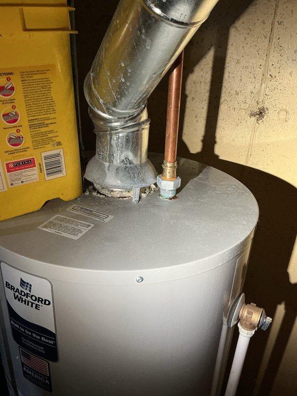 Responded to active water leak in basement originating from area above Bradford White water heater. Upon inspection, identified moisture and condensation on metallic exhaust flue pipe extending from top of water heater unit. Leak had been present for several weeks with recent worsening, contained to immediate vicinity by floor drain. Examined all plumbing connections including copper supply lines with brass fittings and dielectric union, PVC discharge piping, and surrounding utility infrastructure including yellow condensate pump and associated drainage systems. Assessed water heater installation in 1973-era home with concrete block foundation. Documented conditions with photographic evidence showing wet flue pipe, plumbing configuration, and mechanical systems layout. Confined water damage to immediate area around water heater with proper drainage preventing spread throughout basement.