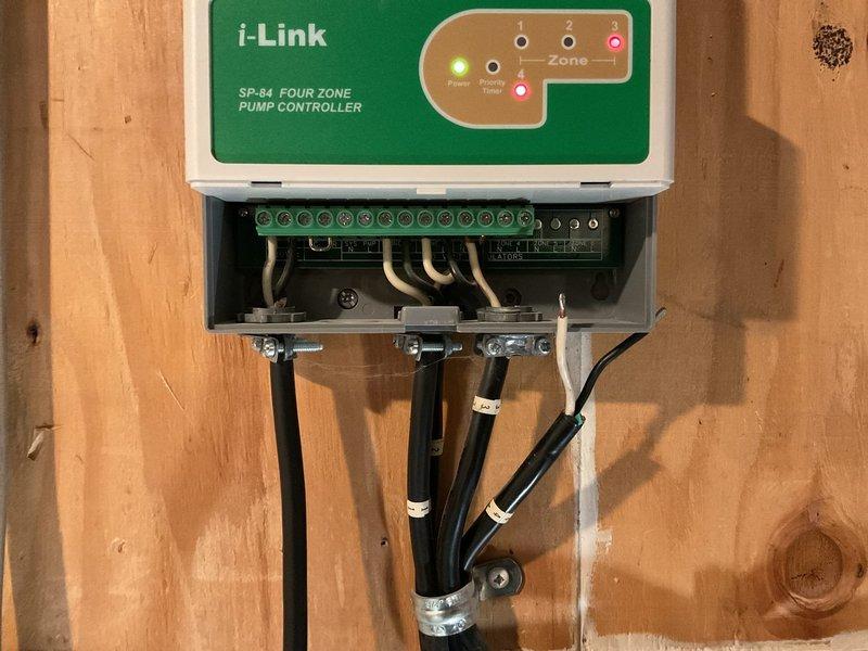 Completed sump pump system replacement and installation of i-Link SP-B4 Four Zone Pump Controller with integrated Grundfos circulator pump. The new controller was mounted to the wall with proper electrical connections made to terminal blocks, featuring multi-zone monitoring capabilities with illuminated power and zone indicator lights. Installed red-housed Grundfos circulator pump on copper piping network with electrical junction box, incorporating brass ball valves and various diameter copper pipe connections for optimal water circulation. All conduits were properly secured with metal clamps and cable ties, and system was tested to verify proper operation across all zones.