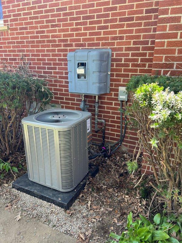 Completed installation of a 2-ton air conditioning system with matching evaporator coil. The outdoor condensing unit was positioned on a rubber isolation pad on the left side of the property against the brick exterior wall, with proper electrical disconnect box mounted above and refrigerant lines and conduit secured to the wall. The indoor air handler was installed in the basement utility area with appropriate drainage piping and electrical connections. All mechanical, electrical, and air conditioning permits were requested and the system was reconnected and tested for proper operation.