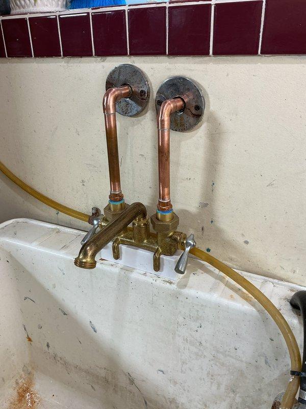 Replaced corroded laundry tub faucet assembly with new basic faucet and shutoff valves. Removed existing copper pipe connections showing extensive green patina and verdigris corrosion on brass fittings and valve mechanisms. Installed new wall-mounted faucet assembly with proper supply line connections to existing copper stub-outs.