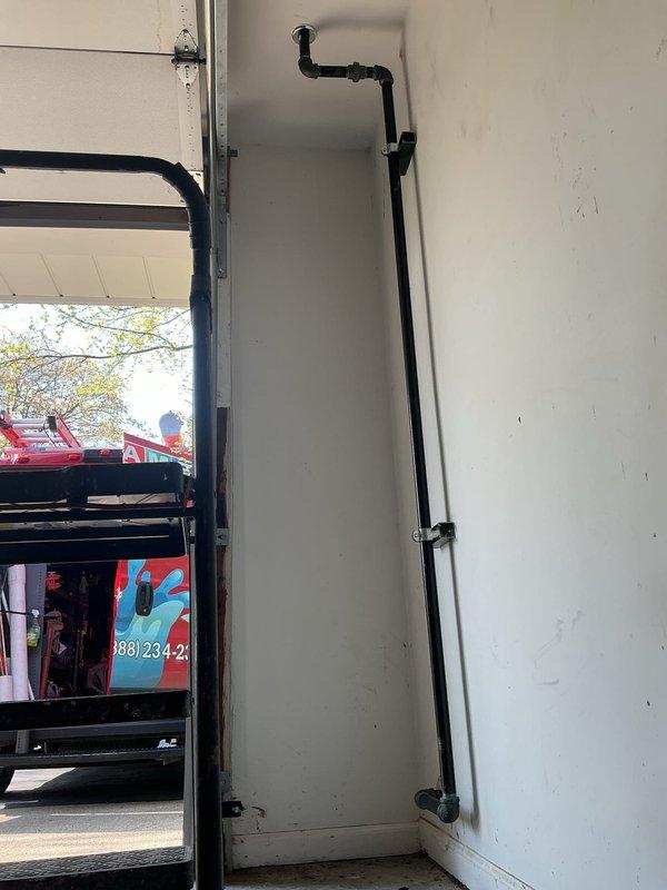 Completed gas line installation in residential property, running black iron pipe vertically along interior corner wall with proper securing brackets at code-compliant intervals. Installation includes multiple fittings and elbows to ensure proper routing and connection. Work performed in utility/laundry area and garage space, with all pipe securely fastened to wall structure using metal straps per industry standards.