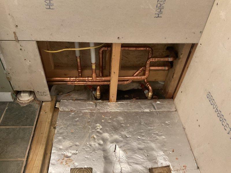 Completed comprehensive plumbing pipe relocation assessment for bathroom renovation project. Evaluated existing copper supply line configuration including two vertical pipes extending from bathroom floor level, plus additional lateral pipes running along wall cavity that extend upward through ceiling structure. Documented extensive network of copper piping with multiple horizontal and vertical runs, brass fittings, T-connections, and elbow joints requiring relocation. Identified white PVC drain lines, yellow flexible gas line connections, and red-handled shut-off valves integrated within existing plumbing assembly. Photographed complete piping layout within exposed wall cavities and crawl space area to provide accurate scope for pipe relocation estimate.
