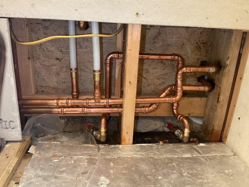 Completed comprehensive plumbing pipe relocation assessment for bathroom renovation project. Evaluated existing copper supply line configuration including two vertical pipes extending from bathroom floor level, plus additional lateral pipes running along wall cavity that extend upward through ceiling structure. Documented extensive network of copper piping with multiple horizontal and vertical runs, brass fittings, T-connections, and elbow joints requiring relocation. Identified white PVC drain lines, yellow flexible gas line connections, and red-handled shut-off valves integrated within existing plumbing assembly. Photographed complete piping layout within exposed wall cavities and crawl space area to provide accurate scope for pipe relocation estimate.