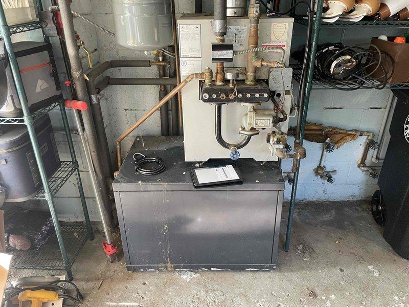 Conducted site assessment for new boiler installation to replace 30-year-old unit located in garage mechanical space. Documented existing system configuration including aged boiler with extensive copper piping, multiple zone valves, expansion tank, and manifold connections. Noted significant water damage and deterioration on interior cabinet wall surfaces, corroded substrate material, and general wear throughout mechanical space indicating end-of-life system conditions requiring complete replacement.