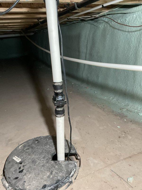 Performed comprehensive inspection of sump pump system located in crawl space. Upon examination, found pump operational with green motor housing positioned in concrete sump pit with standing water present. Noted deteriorated corrugated flexible discharge piping showing significant age-related wear, rust-colored deposits, and discoloration. Inspected white PVC vertical discharge pipe secured with multiple metal clamps along wall, confirming proper installation and structural support. Observed spray foam insulation application on crawl space walls and adequate access for future maintenance. Documented system condition with photographs for service records.