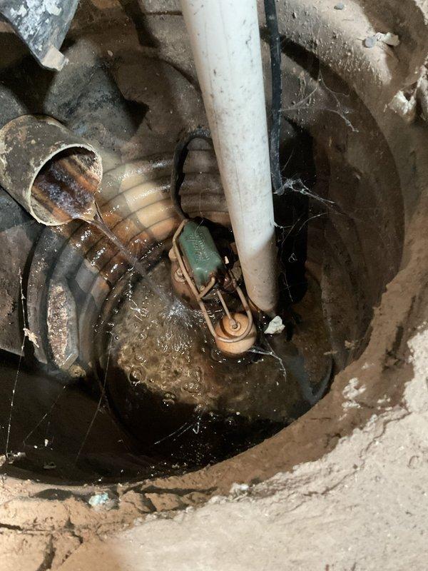 Performed comprehensive inspection of sump pump system located in crawl space. Upon examination, found pump operational with green motor housing positioned in concrete sump pit with standing water present. Noted deteriorated corrugated flexible discharge piping showing significant age-related wear, rust-colored deposits, and discoloration. Inspected white PVC vertical discharge pipe secured with multiple metal clamps along wall, confirming proper installation and structural support. Observed spray foam insulation application on crawl space walls and adequate access for future maintenance. Documented system condition with photographs for service records.