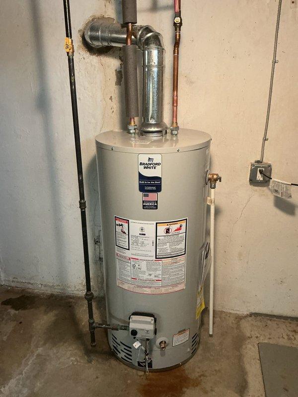 Completed warranty installation of a 40-gallon Bradford White standard tank water heater in residential basement utility area. Reconnected all water supply lines including copper piping and galvanized connections with proper elbow joints, installed pressure relief valve assembly, and connected gas control unit at base of tank. All plumbing connections tested and verified operational with no leaks detected.