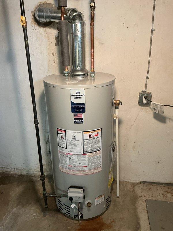 Completed warranty installation of a 40-gallon Bradford White standard tank water heater in residential basement utility area. Reconnected all water supply lines including copper piping and galvanized connections with proper elbow joints, installed pressure relief valve assembly, and connected gas control unit at base of tank. All plumbing connections tested and verified operational with no leaks detected.