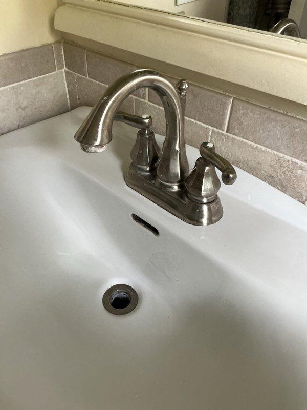Completed bathroom faucet replacement on white porcelain sink basin. Removed existing faucet assembly and installed customer-provided chrome two-handle faucet with curved spout and decorative lever handles. Connected braided flexible supply lines to existing shutoff valves, verified P-trap assembly for proper drainage, tested system for leaks, and confirmed full functionality of hot and cold water flow.