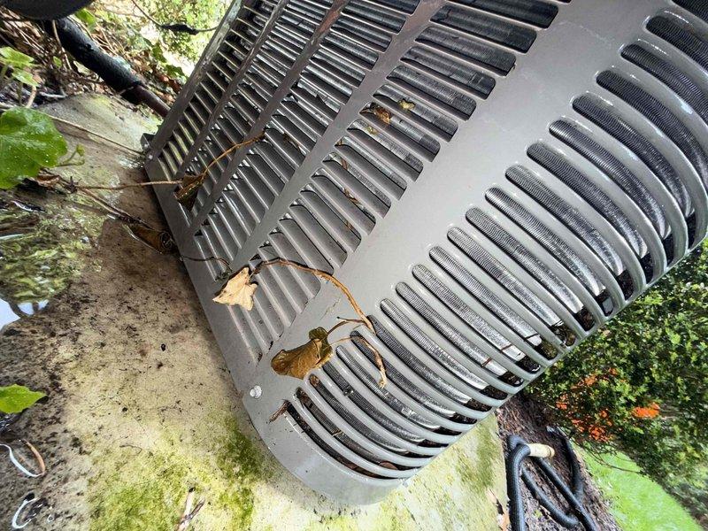 Completed comprehensive Gold AC Inspection on a 10-year-old residential air conditioning system with outdoor condenser unit and basement-located air handler. Performed thorough visual assessment of condenser unit, noting leaf accumulation on louvers and signs of weathering on exterior housing. Inspected refrigerant lines, electrical conduit, and utility connections running from outdoor unit to basement air handler, confirming proper installation and securing of piping. Documented equipment condition with photographs showing condenser placement, surrounding area with moss and algae growth on concrete pad, and exterior utility connections. System confirmed operational at time of inspection, with all components accessible and functioning as designed.