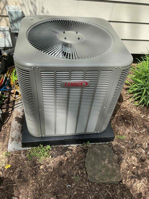 Completed installation of 4-ton Lennox air conditioning condenser unit at rear of residence. Unit positioned on composite platform pad against white vinyl-sided exterior wall with proper clearances maintained for airflow and serviceability. Electrical reconnection performed per code requirements. Mechanical and electrical permits obtained for installation work. System remeasured and verified for proper operation. Condenser features horizontal louvered panels for optimal ventilation and top-mounted circular fan assembly with protective grille. Unit installed on mulched ground surface with adequate drainage and access for future maintenance.