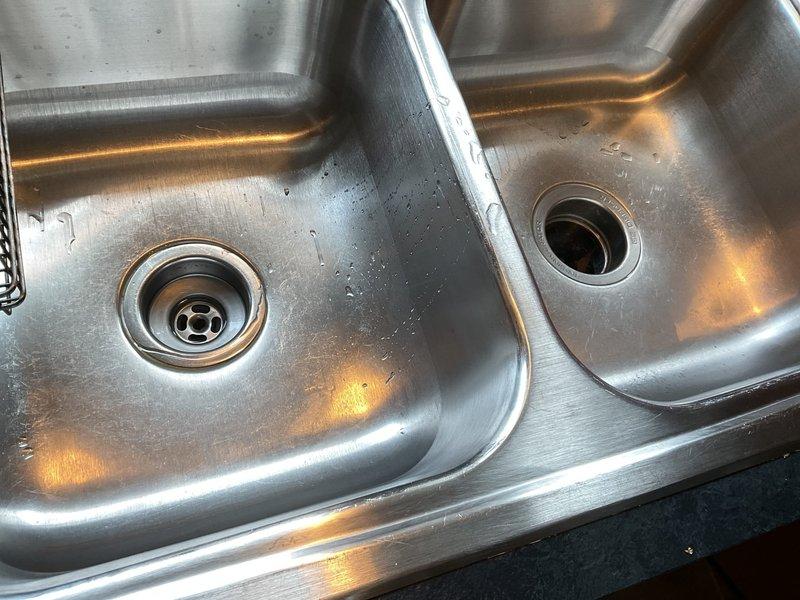 # Clogged Kitchen Drain Service

Responded to service call for backed-up kitchen drain in double-basin stainless steel sink. Upon arrival, performed diagnostic inspection of the drainage system and cabinet plumbing configuration. Inspected P-trap assembly, white PVC drain lines, and garbage disposal unit mounted beneath the sink basin. 

Identified blockage in the drain line causing water backup. Cleared obstruction from the drainage system using appropriate plumbing tools and techniques. Tested both sink basins for proper drainage flow and verified P-trap connections were secure and functioning correctly. Ensured garbage disposal unit was operating properly and all plumbing connections were leak-free. 

Kitchen drain system restored to full functionality with water flowing freely through both basins. All components inspected and confirmed operational before completing service.