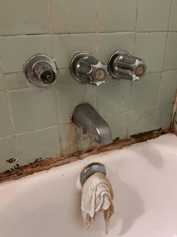 # Bathroom Plumbing Repair and Drain Service

Responded to service call for multiple bathroom plumbing issues including leaking bathtub faucet, broken control knob, and clogged sink drain. Upon inspection, identified deteriorated bathtub fixtures with significant rust staining and water damage along tub-to-wall junction, indicating prolonged moisture exposure.

Accessed concealed plumbing assembly by removing wall cavity access panel, revealing aged copper supply lines with visible corrosion and PVC drain configuration with dual shut-off valves. Addressed clogged sink drain using appropriate clearing methods. Assessed bathtub faucet assembly with three chrome fixtures mounted on tile wall, noting one cross-handle valve required replacement due to mechanical failure. Evaluated extent of rust deterioration at fixture base and tub spout area to determine necessary repairs. Documented existing conditions including compromised sealing at tub-wall interface and aging plumbing infrastructure 