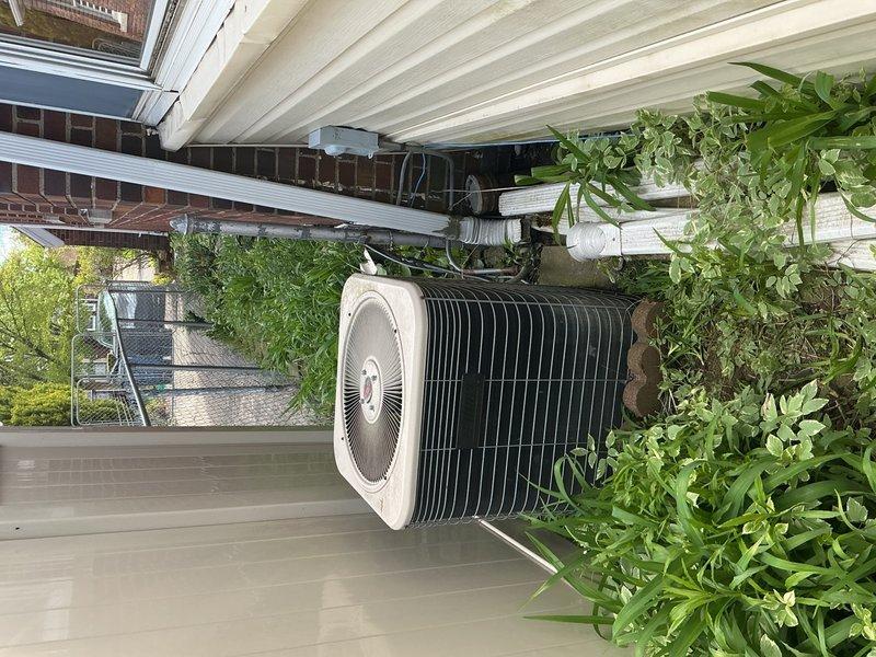 Completed comprehensive Gold AC Inspection on a 20+ year old single-unit air conditioning system with air handler located in basement. Performed thorough visual examination of outdoor condensing unit positioned on elevated deck mounting, inspecting compressor, condenser coil fins, electrical connections, and refrigerant lines. Evaluated electrical wiring connections and terminal blocks for proper voltage and secure terminations. Assessed condensate drainage system and PVC drain lines for proper function. Inspected unit for signs of wear, corrosion, and environmental damage consistent with age of equipment. Verified system operational status and confirmed unit functioning properly at time of inspection. Documented equipment condition through photographic evidence showing exterior condensing unit placement, electrical connections, and surrounding installation area.