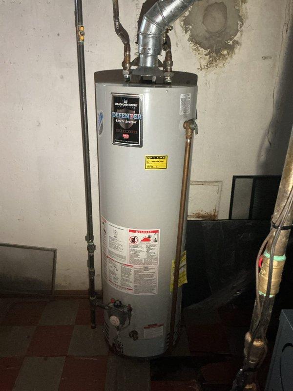 Responded to service call for leaking tanked water heater in basement of 73-year-old residence. Upon inspection, identified a Defense brand residential water heater, approximately 19-21 years old, installed on north basement wall with active leak at tank base. Unit showed signs of significant age and deterioration, with corrosion evident on tank body and compromised structural integrity. Documented existing installation configuration including copper supply lines, metallic exhaust vent system, temperature and pressure relief valve assembly, and gas shutoff valve. Photographed water heater condition, surrounding plumbing connections, ductwork integration, and basement environment showing water damage on adjacent walls. Advised customer that tank failure due to age-related corrosion requires complete water heater replacement, as leaking tank cannot be repaired and poses risk of catastrophic failure and water damage.
