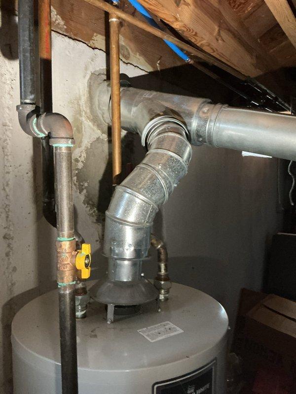 Responded to service call for leaking tanked water heater in basement of 73-year-old residence. Upon inspection, identified a Defense brand residential water heater, approximately 19-21 years old, installed on north basement wall with active leak at tank base. Unit showed signs of significant age and deterioration, with corrosion evident on tank body and compromised structural integrity. Documented existing installation configuration including copper supply lines, metallic exhaust vent system, temperature and pressure relief valve assembly, and gas shutoff valve. Photographed water heater condition, surrounding plumbing connections, ductwork integration, and basement environment showing water damage on adjacent walls. Advised customer that tank failure due to age-related corrosion requires complete water heater replacement, as leaking tank cannot be repaired and poses risk of catastrophic failure and water damage.