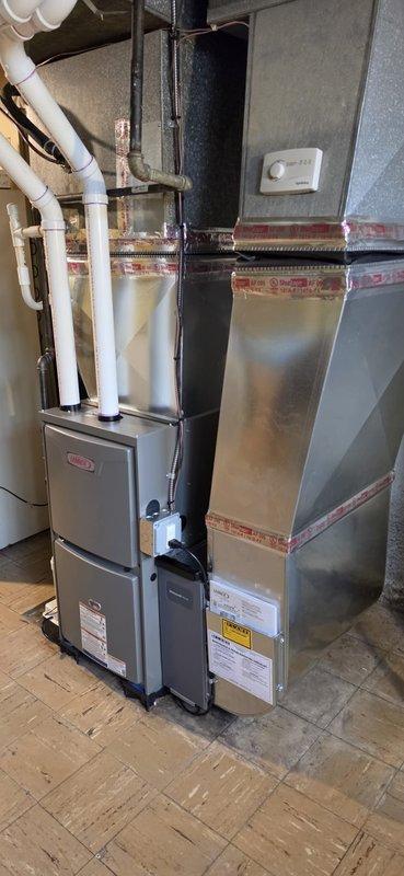 Completed installation of new Lennox residential furnace system in basement utility area. Unit positioned on tiled floor with proper clearances for maintenance access, connected to existing ductwork with black supply plenum extending to ceiling. White PVC vent piping routed and secured for proper exhaust and combustion air intake. Air handler unit installed adjacent to furnace with professional sealing and red tape application along seams. All electrical connections completed, manufacturer labels and safety warnings verified in place. System tested for proper operation, airflow confirmed through duct connections. Permit requested and consumer energy rebate documentation submitted for $100 qualification. Installation meets code requirements with appropriate spacing between components and access to service panels maintained.
