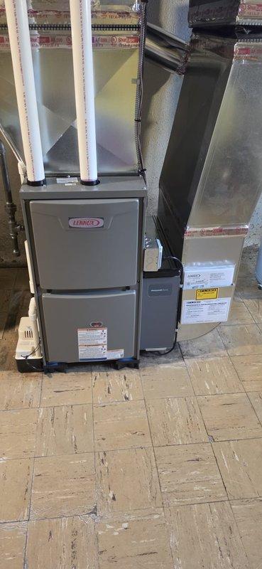 Completed installation of new Lennox residential furnace system in basement utility area. Unit positioned on tiled floor with proper clearances for maintenance access, connected to existing ductwork with black supply plenum extending to ceiling. White PVC vent piping routed and secured for proper exhaust and combustion air intake. Air handler unit installed adjacent to furnace with professional sealing and red tape application along seams. All electrical connections completed, manufacturer labels and safety warnings verified in place. System tested for proper operation, airflow confirmed through duct connections. Permit requested and consumer energy rebate documentation submitted for $100 qualification. Installation meets code requirements with appropriate spacing between components and access to service panels maintained.