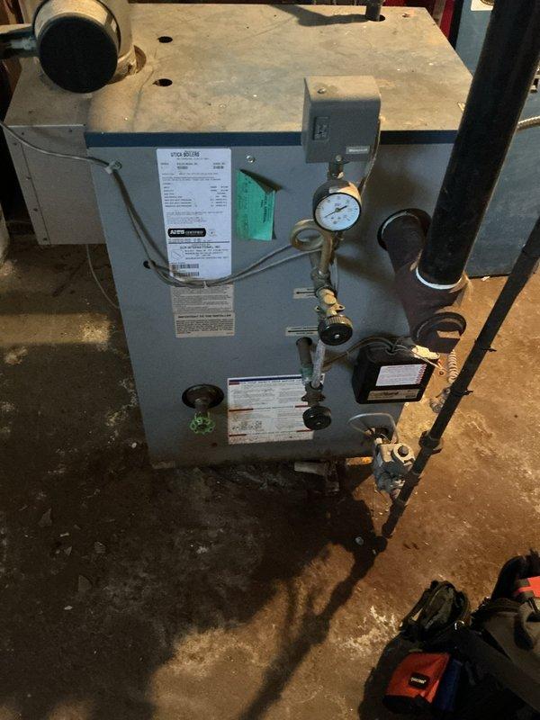 Performed comprehensive tune-up and safety inspection on residential boiler system located in basement. Verified all operational controls, pressure gauge readings, and safety valves including pressure relief valve functionality. Inspected combustion chamber, heat exchanger surfaces, and venting system for proper operation and signs of deterioration. Checked all piping connections, brass fittings, and isolation valves for leaks or corrosion. Examined electrical components and junction box connections for secure mounting and proper wiring. Noted significant rust and corrosion present on secondary piping in crawlspace area beneath unit, with evidence of moisture exposure and deteriorated conditions in surrounding utility space. Documented concrete block foundation showing water staining and aging, with exposed soil floor area containing debris accumulation. System currently operational with all primary safety controls functioning within normal parameters, though crawlspace conditions indi