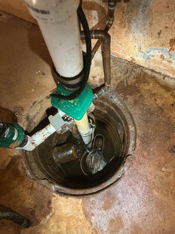 Responded to service call for loose sump pump piping and garbage disposal inspection. Upon arrival, conducted thorough assessment of sump pump system installed in basement floor pit. Inspected submersible pump assembly, discharge piping, and check valve components. Noted existing installation featuring white PVC discharge pipe with green check valve fittings and secondary inlet piping. Examined pump control float switch mechanism with visible corrosion on metal components and connections. Evaluated structural integrity of piping connections and mounting hardware. Observed aged system with moisture exposure, rust accumulation on metal fittings, and general operational wear throughout assembly. Checked electrical connections and wiring for proper function and safety compliance. Assessed concrete sump pit condition and surrounding floor area showing moisture staining and deterioration. Inspected galvanized steel piping components and verified check valve operation to prevent backflow. Eva