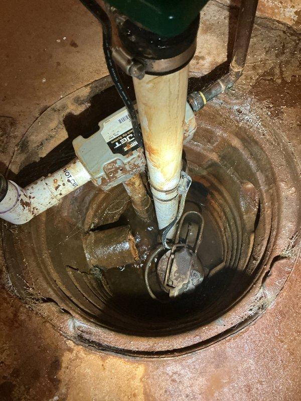 Responded to service call for loose sump pump piping and garbage disposal inspection. Upon arrival, conducted thorough assessment of sump pump system installed in basement floor pit. Inspected submersible pump assembly, discharge piping, and check valve components. Noted existing installation featuring white PVC discharge pipe with green check valve fittings and secondary inlet piping. Examined pump control float switch mechanism with visible corrosion on metal components and connections. Evaluated structural integrity of piping connections and mounting hardware. Observed aged system with moisture exposure, rust accumulation on metal fittings, and general operational wear throughout assembly. Checked electrical connections and wiring for proper function and safety compliance. Assessed concrete sump pit condition and surrounding floor area showing moisture staining and deterioration. Inspected galvanized steel piping components and verified check valve operation to prevent backflow. Eva