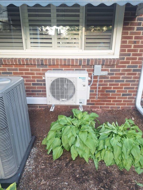 Completed installation of Daikin mini-split system in rear yard area of residential property. Mounted outdoor condenser unit on brick exterior wall using white mounting brackets and installed indoor console unit. Ran new dedicated electrical circuit to support the system, with mechanical, electrical, and mini-split permits requested and processed. Unit positioned on concrete pad in landscaped area, with all refrigerant lines and electrical connections properly secured and tested for optimal operation.