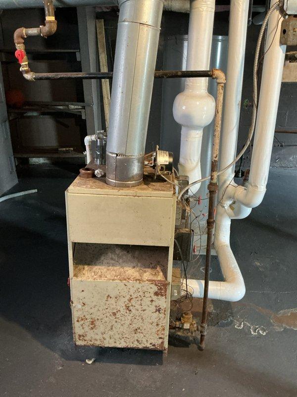 Conducted estimate for steam boiler system in basement showing evidence of flooding and water damage. Equipment operational at time of inspection but displaying significant corrosion and rust deterioration on cabinet housing, particularly at base and lower panels. Water staining patterns visible on floor surrounding unit indicate previous or ongoing moisture intrusion issues. Piping network includes copper supply lines with brass fittings showing oxidation, white PVC venting, expansion tank, and flexible metal conduit connections. System age estimated at 10+ years with visible wear throughout. Documented extensive rust damage to boiler cabinet exterior and corrosion on copper piping. Photographed overall system condition including deteriorated metal surfaces, complex piping configuration, and floor water damage patterns for estimate preparation. Equipment currently functioning despite compromised cabinet integrity and moisture exposure history.