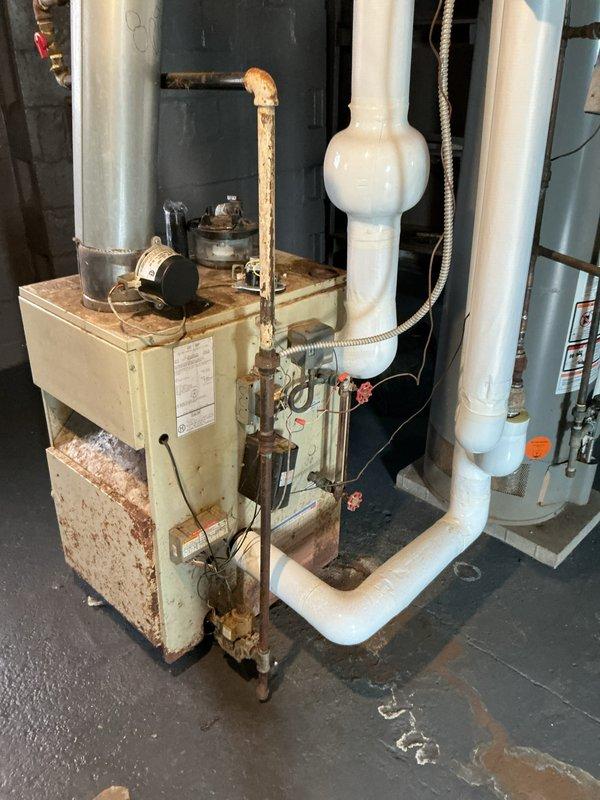 Conducted estimate for steam boiler system in basement showing evidence of flooding and water damage. Equipment operational at time of inspection but displaying significant corrosion and rust deterioration on cabinet housing, particularly at base and lower panels. Water staining patterns visible on floor surrounding unit indicate previous or ongoing moisture intrusion issues. Piping network includes copper supply lines with brass fittings showing oxidation, white PVC venting, expansion tank, and flexible metal conduit connections. System age estimated at 10+ years with visible wear throughout. Documented extensive rust damage to boiler cabinet exterior and corrosion on copper piping. Photographed overall system condition including deteriorated metal surfaces, complex piping configuration, and floor water damage patterns for estimate preparation. Equipment currently functioning despite compromised cabinet integrity and moisture exposure history.