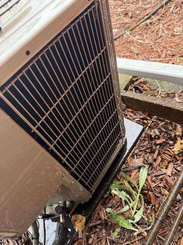 Completed comprehensive Gold AC inspection on residential cooling system less than 10 years old with air handler located in basement. Performed thorough examination of outdoor condenser unit, which showed normal weathering and debris accumulation on exterior grilles and fan guard. Inspected refrigerant line connections, service valves, and unit mounting on concrete pad with plastic base. Evaluated condenser coil condition through louvers, noting dirt and debris buildup between slats requiring cleaning for optimal airflow and efficiency. Assessed fan assembly operation and verified proper clearance around unit, though landscaping encroachment with mulch, leaves, and vegetation was observed near foundation and base perimeter. Documented unit specifications and current operational status through photographic evidence. System confirmed operational at time of inspection.