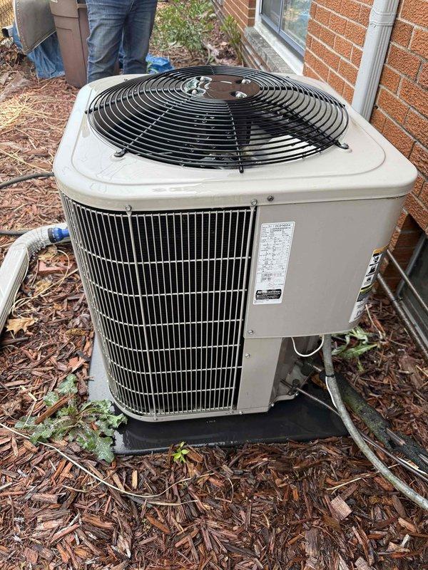 Completed comprehensive Gold AC inspection on residential cooling system less than 10 years old with air handler located in basement. Performed thorough examination of outdoor condenser unit, which showed normal weathering and debris accumulation on exterior grilles and fan guard. Inspected refrigerant line connections, service valves, and unit mounting on concrete pad with plastic base. Evaluated condenser coil condition through louvers, noting dirt and debris buildup between slats requiring cleaning for optimal airflow and efficiency. Assessed fan assembly operation and verified proper clearance around unit, though landscaping encroachment with mulch, leaves, and vegetation was observed near foundation and base perimeter. Documented unit specifications and current operational status through photographic evidence. System confirmed operational at time of inspection.
