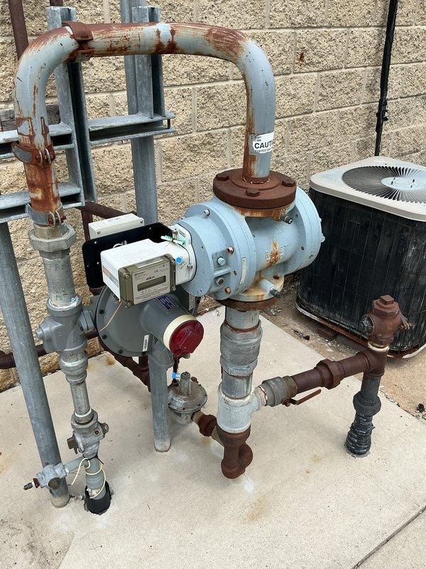 Conducted comprehensive site assessment for gas line installation serving outdoor mechanical equipment. Evaluated existing corroded piping infrastructure including blue-painted pump assembly with deteriorated flanges and oxidized connections, documented current system configuration with digital meter and pressure gauge components mounted on concrete pad adjacent to HVAC unit. Assessed backyard routing requirements from utility connection point through lawn area to equipment location, identified excavation pathway and measured distances for gas line extension to service mechanical systems at rear of residential structure.